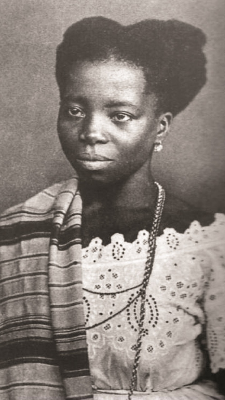 Retrato antigo em preto e branco de Tia Ciata. Ela é uma mulher negra de meia-idade, com um penteado alto e volumoso (coque ou turbante). Ela veste um vestido claro com decote rendado ou bordado e um longo colar de contas. Um xale ou manta listrada está drapeado sobre seu ombro esquerdo. Seu olhar é direto e sério.