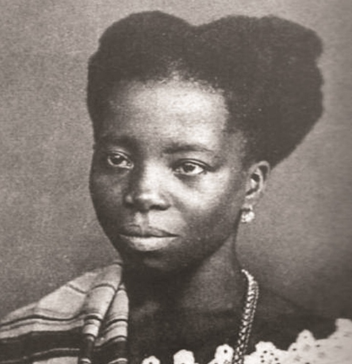 Retrato antigo em preto e branco de Tia Ciata. Ela é uma mulher negra de meia-idade, com um penteado alto e volumoso (coque ou turbante). Ela veste um vestido claro com decote rendado ou bordado e um longo colar de contas. Um xale ou manta listrada está drapeado sobre seu ombro esquerdo. Seu olhar é direto e sério.