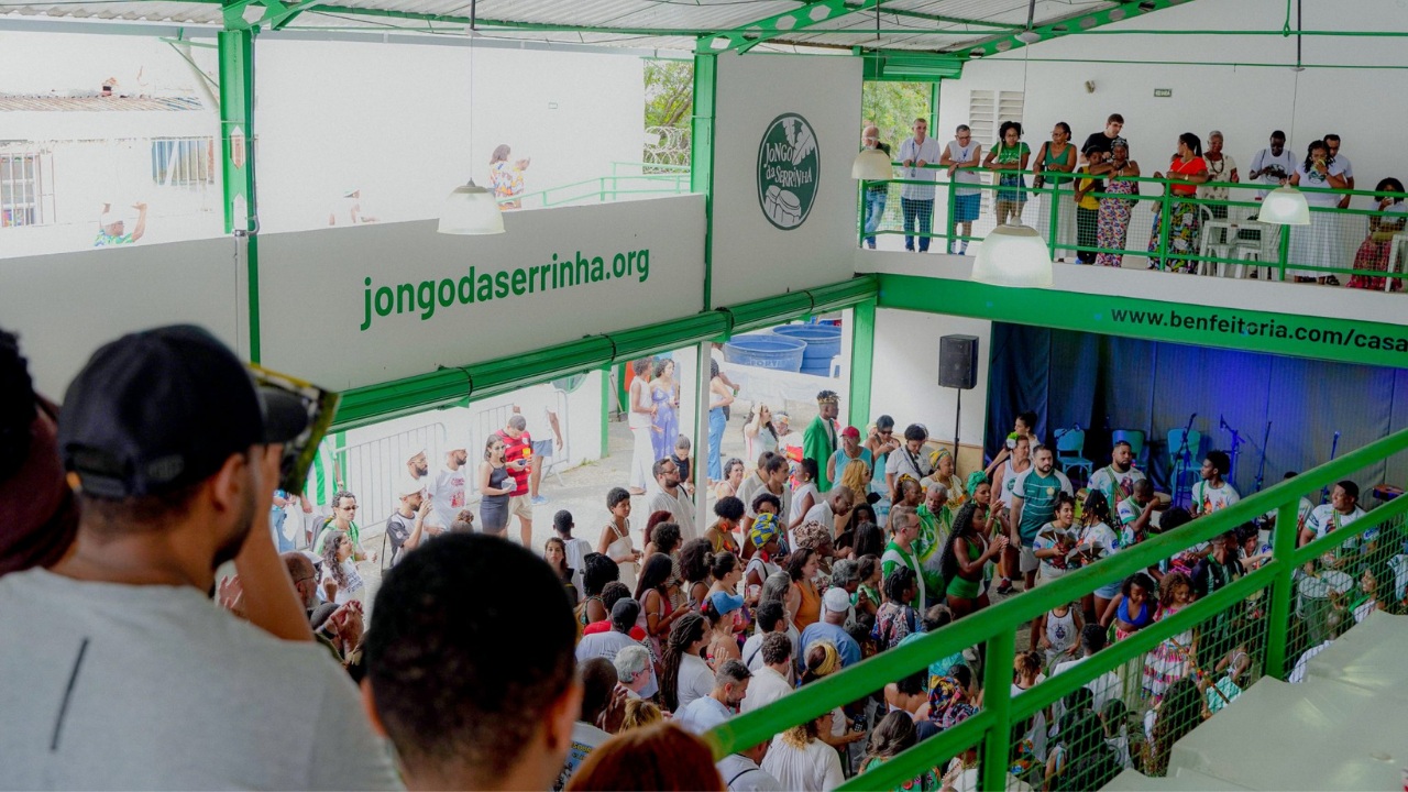 Multidão reunida em um espaço coberto, de estrutura verde e branca, possivelmente o local de um evento do Jongo da Serrinha. No nível inferior, um grande grupo de pessoas está reunido e interagindo, algumas vestindo roupas brancas e verdes. No nível superior, há uma varanda onde outras pessoas observam a cena. O logotipo "Jongo da Serrinha" e o endereço "jongodaserrinha.org" estão visíveis nas estruturas.