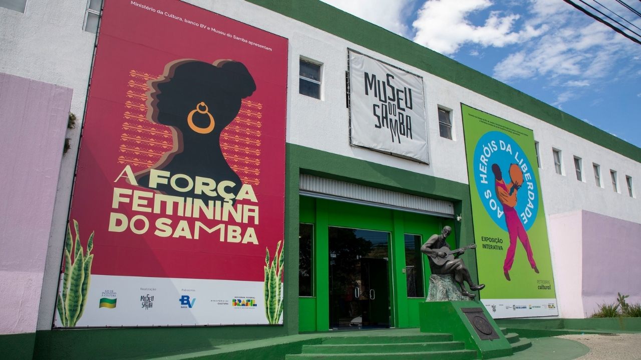 Fachada do Museu do Samba, no Rio de Janeiro, com duas grandes faixas de exposição: à esquerda, um cartaz vermelho com o título “A Força Feminina do Samba” e a silhueta de uma mulher com brinco dourado; à direita, um cartaz verde e azul da exposição “Os Heróis da Liberdade”, com duas figuras dançando. Em frente à entrada verde, há uma escultura de um músico tocando violão.