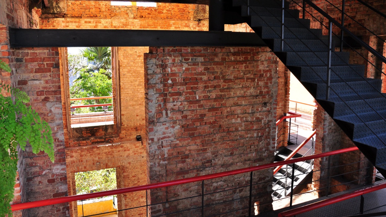 Vista interna de um edifício antigo, provavelmente em ruínas, com paredes de tijolos vermelhos expostos. Uma escada moderna em metal preto sobe diagonalmente no canto direito. Há vigas de metal preto sustentando a estrutura e corrimãos vermelhos brilhantes em várias varandas internas. Através das janelas abertas, vê-se vegetação verde e luz natural.