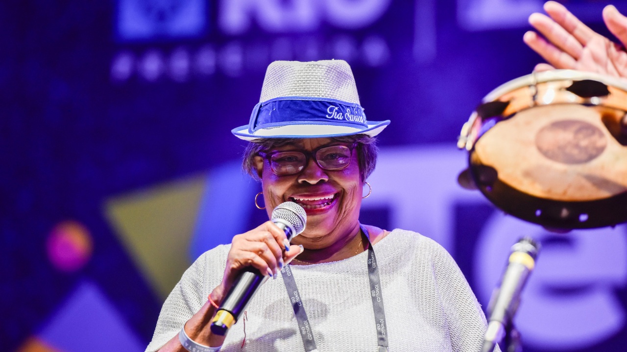 Mulher mais velha sorrindo enquanto canta em um microfone, usando chapéu azul e branco, óculos e blusa clara. Ela está em um palco iluminado por luzes coloridas, com um pandeiro parcialmente visível ao lado.
