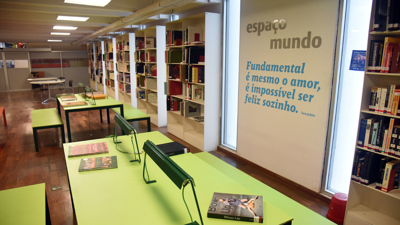 Ambiente interno da Biblioteca Parque com iluminação clara e organizada. Em primeiro plano, mesas retangulares verde-claro com luminárias individuais verdes, sobre as quais estão dispostos livros e revistas. Ao redor, estantes brancas altas repletas de livros formam corredores de leitura. No fundo, há mesas e cadeiras para estudo. À direita, uma parede clara exibe o texto “espaço mundo” e a frase em azul: “Fundamental é mesmo o amor, é impossível ser feliz sozinho”, atribuída a Tom Jobim, próxima a grandes janelas que deixam entrar luz natural.