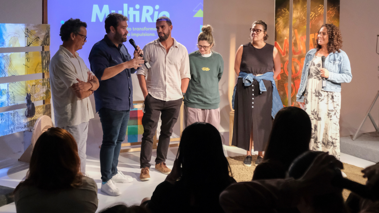 **Texto alternativo (alt text):** Seis pessoas adultas estão em pé sobre um palco, voltadas para o público, durante um evento. Uma delas segura um microfone e fala, enquanto as outras escutam atentamente. Ao fundo, há uma tela com a palavra “MultiRio” e elementos visuais coloridos, além de painéis decorativos. Na parte inferior da imagem, vê-se a silhueta do público sentado, indicando um momento de conversa ou apresentação coletiva em um ambiente cultural ou educacional.