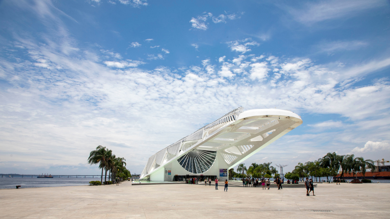 Vista externa do Museu do Amanhã em dia ensolarado, com céu azul e nuvens espalhadas. A construção branca e futurista, marcada por linhas alongadas e estrutura metálica aparente, se projeta sobre a praça ampla em frente à Baía de Guanabara. Algumas pessoas caminham pelo espaço aberto ao redor do edifício, enquanto palmeiras e elementos da orla completam a paisagem.