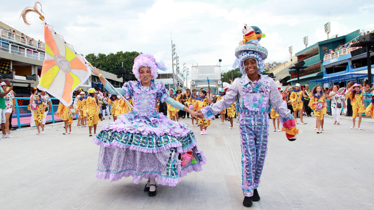 Duas crianças da escola de samba mirim Corações Unidos do CIEP desfilam de mãos dadas no centro da avenida, sorrindo enquanto avançam em direção à câmera.  A criança à esquerda usa um vestido volumoso em tons de lilás, roxo e verde-água, com várias camadas de babados, rendas e aplicações brilhantes. A saia é ampla e rodada, formando um círculo estruturado ao redor do corpo. O corpete é ricamente decorado com flores e detalhes em relevo. As mangas são ajustadas e azul-claras, e a maquiagem artística é colorida e marcante, destacando os olhos com desenhos vibrantes. Na cabeça, há uma peruca grande, encaracolada e lilás. Ela segura uma bandeira com segmentos em amarelo, branco e laranja, presa a um mastro fino inclinado para o lado.  A criança à direita veste um conjunto lilás e azul com estampa quadriculada e aplicações florais. O figurino inclui calça comprida e blusa de mangas longas com botões e enfeites decorativos. Na cabeça, usa um chapéu grande e ornamentado, em camadas circulares, com um boneco decorativo no topo. Ela sorri amplamente enquanto segura um pequeno adereço colorido na mão livre.  Ao fundo, outras crianças da escola desfilam organizadas em alas, muitas com fantasias amarelas estampadas e segurando adereços circulares. Nas laterais da avenida há arquibancadas com público e estruturas metálicas. O céu está parcialmente nublado, e a iluminação suave realça as cores vibrantes das fantasias e o clima festivo do desfile.