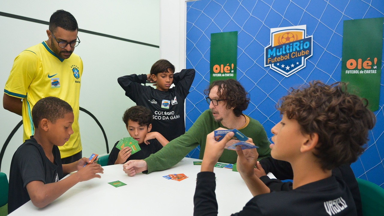 Fotografia. Quatro crianças e um adulto em volta de uma mesa jogando um jogo de cartas.