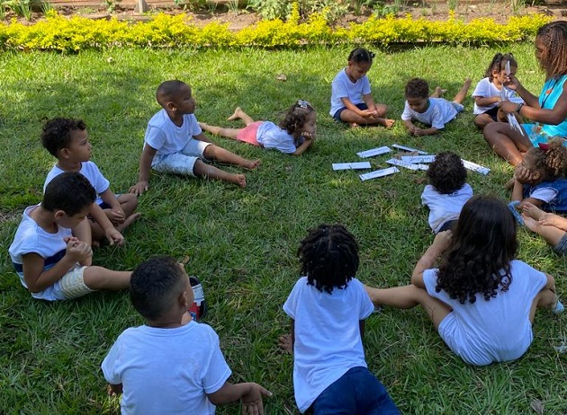 Foto. Crianças pequenas estão em roda com uma professora sobre a grama ao ar livre. Há fichas com os nomes delas no chão. A professora manuseia algumas dessas fichas.