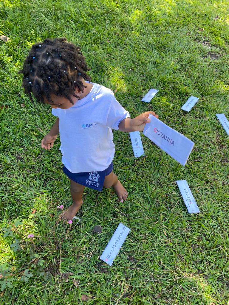 Foto. Menina pequena carrega ficha na qual está escrito Giovanna. Ela veste uniforme escolar, tem os cabelos e pele escuros e caminha sobre a grama.