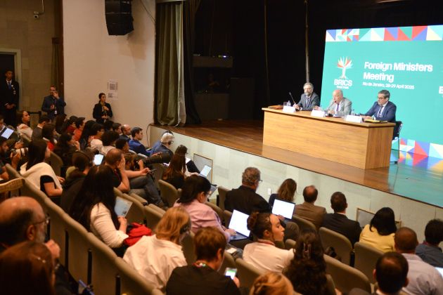 Foto. Auditório com pessoas na plateia sentadas com celulares e notebooks na mão assistem a 3 homens no palco dando entrevista coletiva de imprensa.o 
