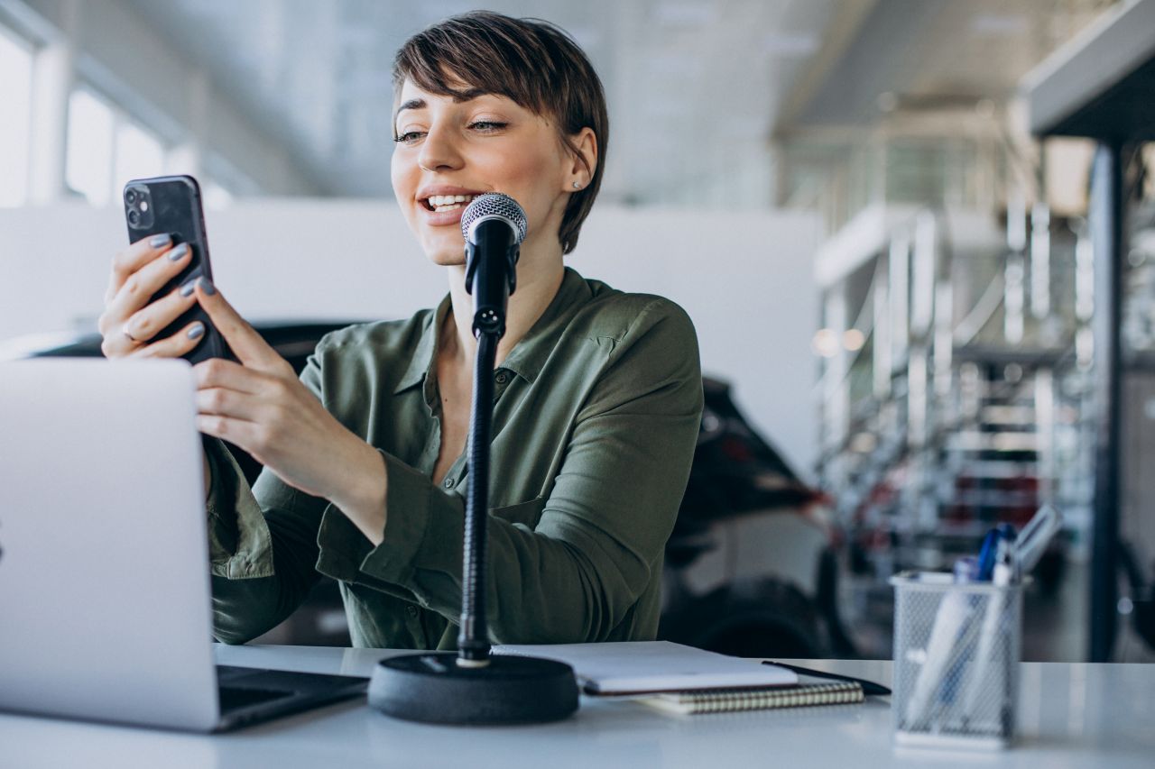 Jornalista com celular na mão apoiada em mesa com notebook e microfone