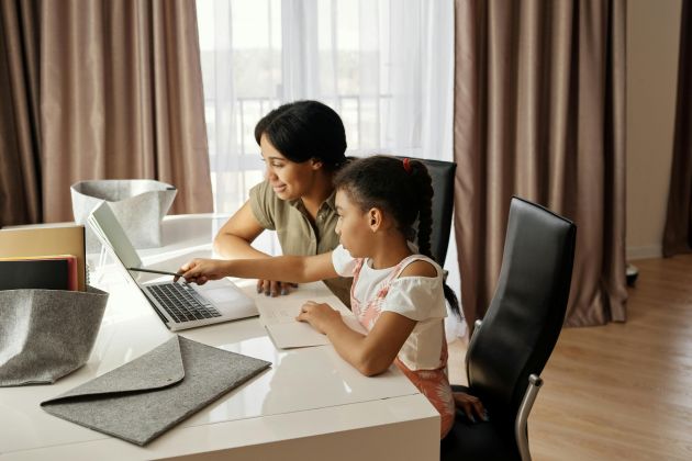 Foto. Uma mulher e uma menina estão sentadas à mesa em frente a um computador portátil em uma mesa branca em uma sala, com janelas ao fundo e cortinas marrons. A mulher está inclinada para a frente, olhando atenta para o computador, e a menina está sentada ligeiramente para trás, apontando para a tela do computador com a mão.  A atmosfera da imagem é de interação familiar entre mãe e filha e concentração.