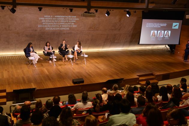  Foto. Quatro mulheres sentadas em cadeiras em um palco. Plateia na parte inferior. A direita, telão com projeção da apresentação.