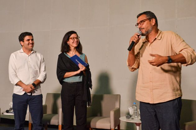 Foto. Um homem com microfone na mão está falando; um homem e uma mulher ao lado dele, também com microfones em mãos, o olham e sorriem.