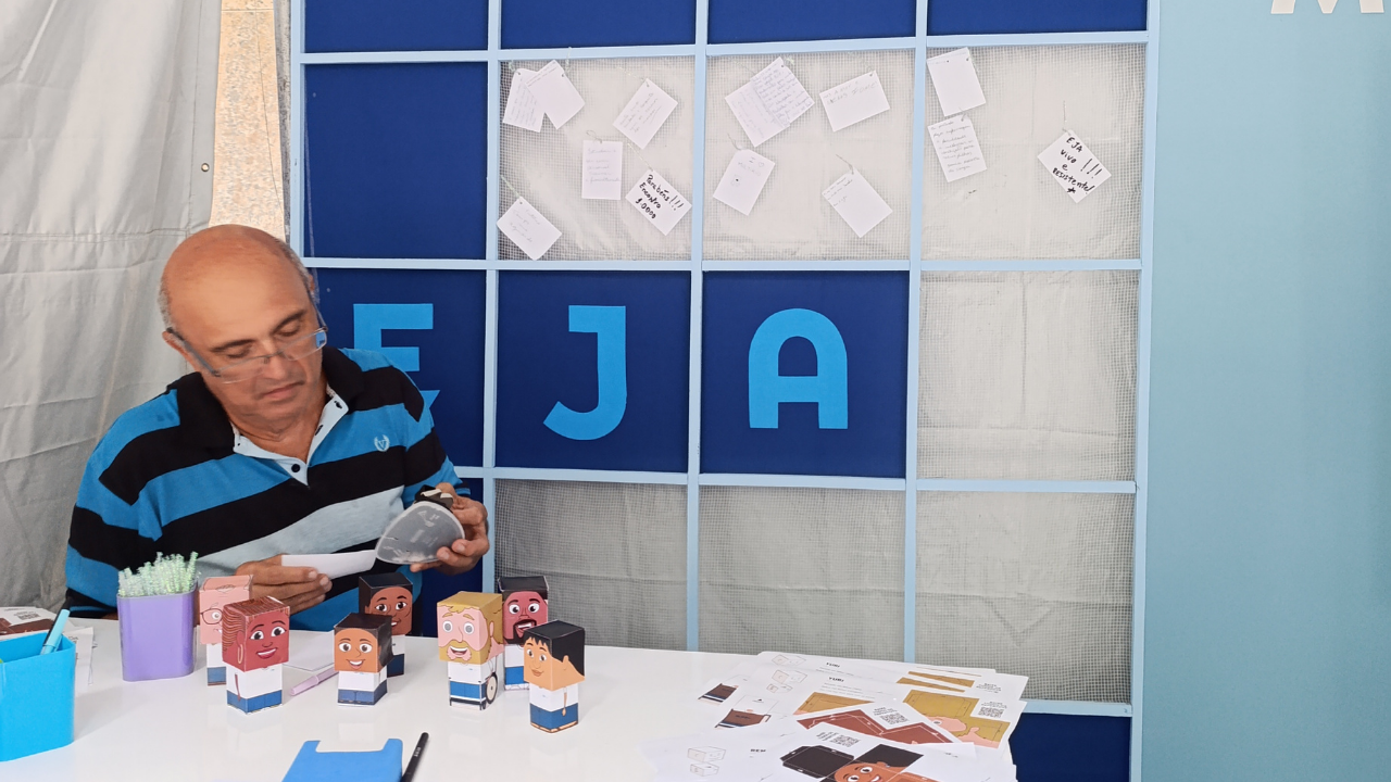 Um homem adulto sentado à mesa, em um espaço de evento da EJA. Ele usa camisa polo listrada em preto e azul e manuseia materiais de papel para montar bonecos tridimensionais. Sobre a mesa há vários bonecos coloridos já montados, canetas, papéis e recipientes com materiais de artesanato. Ao fundo, uma estrutura quadriculada azul exibe letras que formam “EJA” e pequenos bilhetes pendurados.