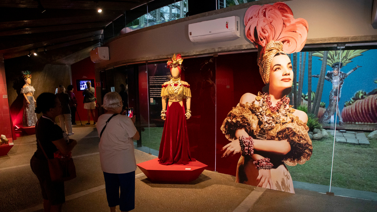 Interior do Museu Carmen Miranda, com iluminação baixa e ambientação em tons avermelhados. Em primeiro plano, visitantes observam figurinos e elementos relacionados à trajetória artística da cantora e atriz Carmen Miranda. No centro da imagem, um manequim sem rosto exibe um vestido longo vermelho com detalhes dourados, colares, pulseiras e um elaborado adorno de cabeça inspirado nos figurinos usados pela artista. À direita, há uma grande imagem de Carmen Miranda usando traje luxuoso com plumas rosas, joias e acessórios dourados, sorrindo levemente e com os braços cruzados. Ao fundo, outros figurinos brilhantes estão expostos em pedestais iluminados, enquanto algumas pessoas circulam pelo espaço observando a exposição. O ambiente possui teto de madeira escura, paredes vermelhas e amplas janelas de vidro laterais, que deixam entrar luz natural.