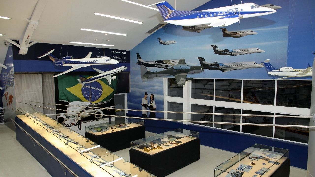 Interior de um espaço expositivo dedicado à aviação, com diversas maquetes de aeronaves suspensas no teto e expostas em vitrines. As paredes são decoradas com imagens de aviões em voo e uma bandeira do Brasil ao fundo. No centro da sala, há painéis com objetos e documentos relacionados ao setor aeroespacial. O ambiente é iluminado por luzes artificiais e apresenta uma organização voltada para a história e a tecnologia da aviação.