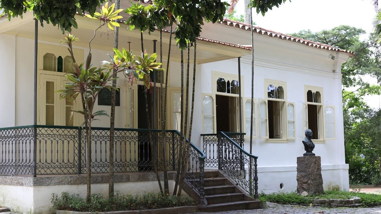 Casa histórica térrea de fachada branca, com janelas amplas e varanda cercada por gradis ornamentados de ferro. Uma escadaria central dá acesso à entrada principal. O imóvel está rodeado por árvores e vegetação, que projetam sombras sobre a construção. À direita da escadaria, há um busto sobre um pedestal de pedra. O ambiente transmite a atmosfera de um espaço cultural ou museu inserido em uma área arborizada.