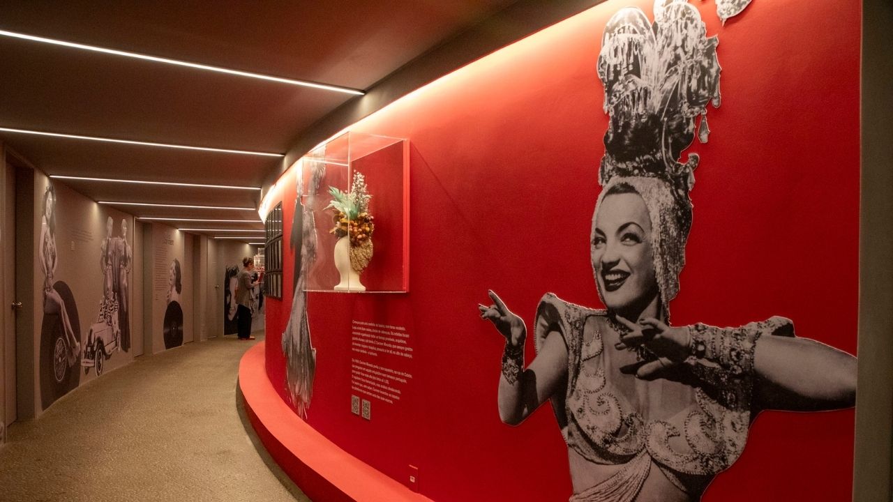 Fotografia do corredor curvo de uma exposição, tirada em ângulo aberto e médio, com o chão de cimento e o teto rebaixado com fitas de LED brancas. A parede da direita é pintada de vermelho vibrante, que traz, em destaque, uma colagem fotográfica da Carmem Miranda, em preto e branco, na qual ela sorri olhando para fora da imagem, com as mãos abertas próximas ao queixo. O resto da colagem tem sua silhueta de perfil, em pé. Do lado esquerdo de sua cabeça, tem uma vitrine transparente em 3D, em formato retangular, contendo um turbante branco de base com diversos adereços de tecidos brancos, dourados e pedrarias. A parede à esquerda é texturizada de cinza-claro, com outras fotos em preto e branco. Ao fundo do corredor, do lado esquerdo, avista-se, de longe, uma pessoa em pé, de casaco longo azul-claro, camisa cinza e calça preta.