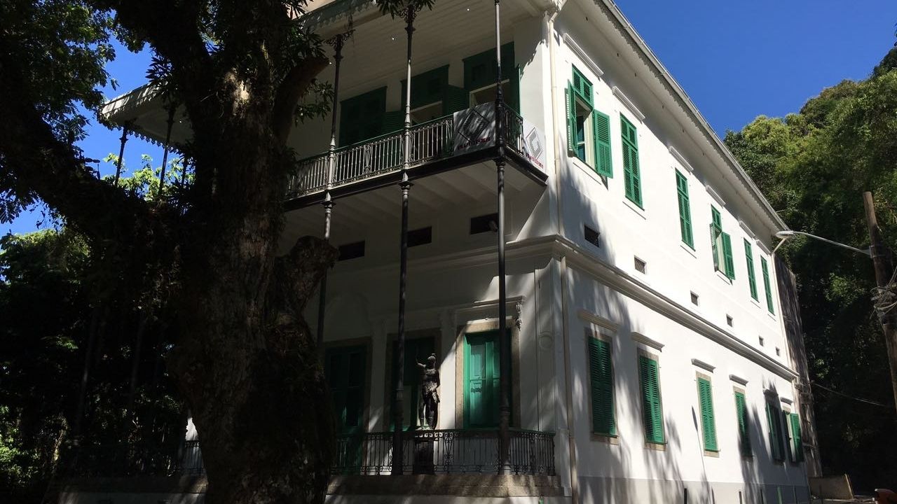 Casarão histórico de dois andares, com fachada branca e janelas de madeira pintadas de verde. O prédio possui uma varanda no pavimento superior, sustentada por colunas metálicas ornamentadas. À frente, uma grande árvore projeta sombras sobre a construção, cercada por vegetação abundante. O céu está limpo e azul, destacando o contraste entre a arquitetura e a paisagem arborizada ao redor.