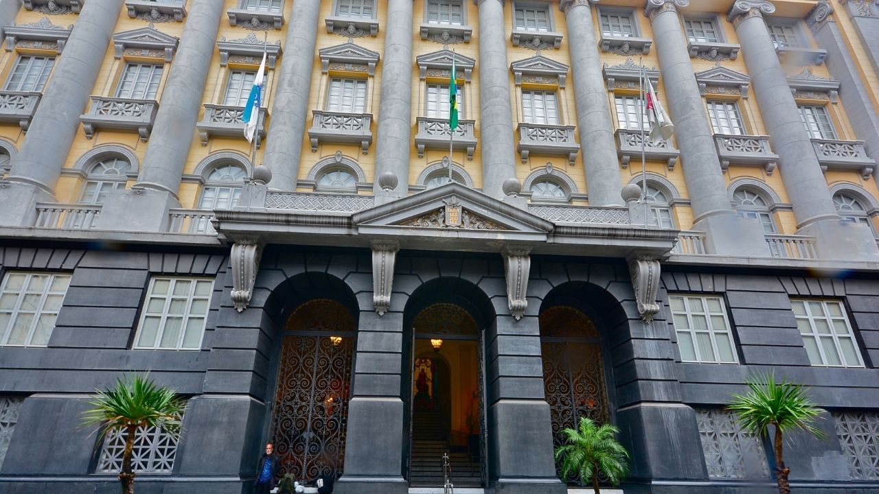 Fachada de um edifício histórico de estilo neoclássico, com paredes em tons de amarelo e cinza, grandes colunas verticais e janelas simétricas distribuídas em vários andares. Na entrada principal, três arcos dão acesso ao interior do prédio, com portões ornamentados em ferro. Bandeiras estão hasteadas acima da entrada, e pequenas palmeiras e plantas decoram a área externa. Uma pessoa aparece próxima à porta, destacando a imponência da construção.