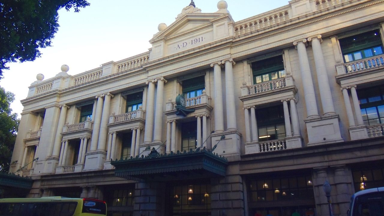 Fachada de um edifício histórico de arquitetura neoclássica, com grandes colunas, sacadas ornamentadas e detalhes em pedra clara. No topo da construção, está inscrito o ano “A.D. 1911”. A entrada principal possui uma marquise decorada, enquanto árvores emolduram parte da cena. Na rua, aparecem parcialmente dois ônibus em frente ao prédio, sob um céu claro.