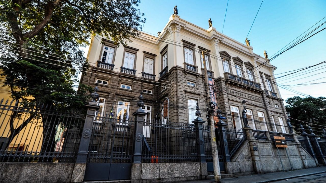 Fotografia em perspectiva de um edifício histórico de três pavimentos, com arquitetura neoclássica e fachada imponente nas cores cinza e creme. A construção ocupa a maior parte da imagem e é vista a partir do nível da rua, de baixo para cima, destacando sua grandiosidade. O piso térreo e parte do primeiro andar apresentam revestimento em pedra escura trabalhada, enquanto o andar superior possui acabamento claro, com molduras ornamentadas ao redor das janelas e detalhes decorativos na cobertura.  Diversas janelas altas e estreitas, com esquadrias brancas e pequenas sacadas de ferro preto, se distribuem simetricamente pela fachada. No topo do edifício, há esculturas posicionadas sobre o coroamento da construção. Em frente ao prédio, um gradil metálico preto de grande porte cerca toda a propriedade. Escadarias de pedra conduzem às entradas laterais.  Uma árvore de copa ampla ocupa parte do lado esquerdo da imagem, projetando sombra sobre a fachada. Vários fios e cabos elétricos atravessam a cena horizontal e diagonalmente, passando à frente do prédio. O céu está limpo e azul, sugerindo um dia ensolarado. A rua em primeiro plano está vazia, reforçando o destaque visual da construção histórica. Na fachada é possível ler a inscrição “Lar José de Mello”, indicando o nome do edifício.